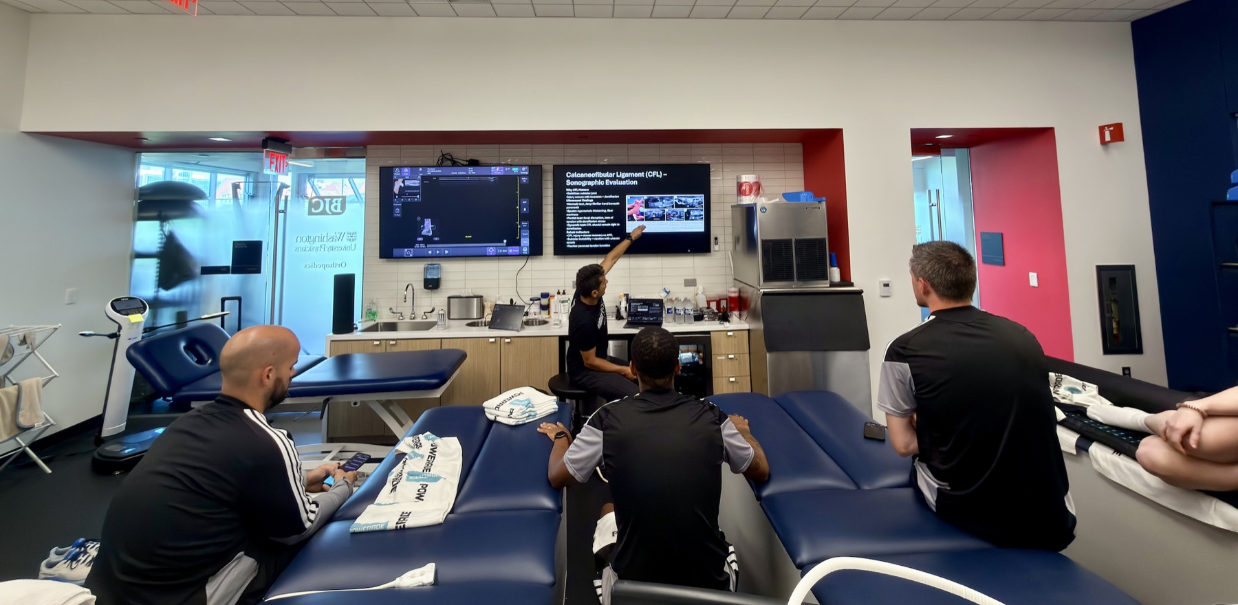 A Pura Health instructor explaining ankle ultrasound images to a group of clinicians in a team treatment room